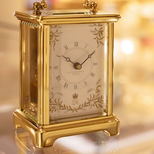 Gold carriage clock with a gold handle and black presentation box.  There is a clock face surrounded by the design of wheat ears and olive branches. On the clock face are the words 'Platinum Jubilee 2022' and an ER surmounted by the crown.