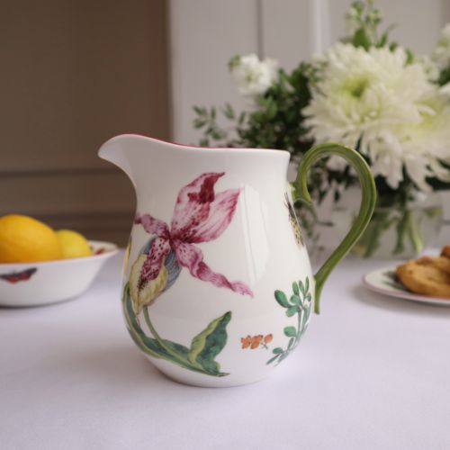 Milk jug on table cloth with bowl of lemons and flowers in background. 