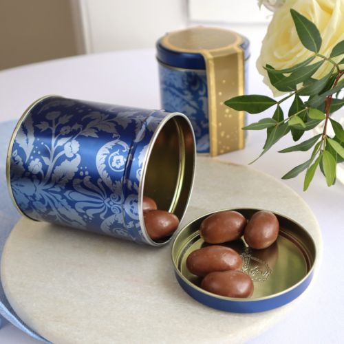 Blue tin with acanthus print and crown embellished lid. A pile of chocolate covered brazil nuts next to the tin. 